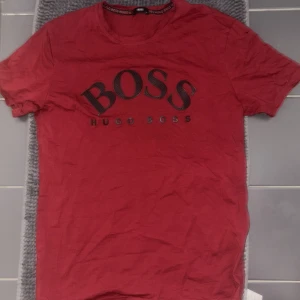 Röd t-shirt från Hugo Boss - Autentisk röd t-shirt från Hugo Boss med stort BOSS-tryck på framsidan. Det är L men känns som M. Perfekt för en stilren look. Tillverkad i mjukt material för extra komfort.