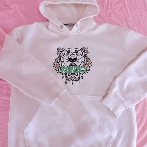 Vit hoodie från Kenzo med tigerbroderi - Säljer en snygg vit hoodie från Kenzo med ett coolt tigerbroderi i svart och grönt på framsidan. Hoodien har en klassisk passform med känguruficka och ribbade muddar vid ärmslut och nederkant. Perfekt för en stilren look!