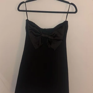 Svart klänning med rosett - Elegant svart klänning med en stor rosett framtill. Axelbandslös design som ger en stilren look. Perfekt för en kväll ute eller en speciell tillställning. Storleken är S-M. Aldrig använd pga för liten.