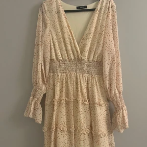 Prickig klänning från Shein - Charmig prickig klänning från Shein i ljus färg med små röda prickar. Klänningen har en v-ringad design och långa ärmar med volangdetaljer. Den smockade midjan ger en smickrande passform och kjolen har flera lager med volanger.