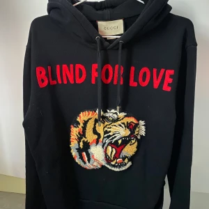 Svart blind in love hoodie från Gucci med tigerbroderi - Säljer en svart hoodie från Gucci med texten 'BLIND FOR LOVE' i rött och ett detaljerat tigerbroderi på framsidan. Hoodien har en klassisk passform, perfekt för vilken dag som helst. I princip helt oanvänd på grund av för liten storlek för mig själv. 