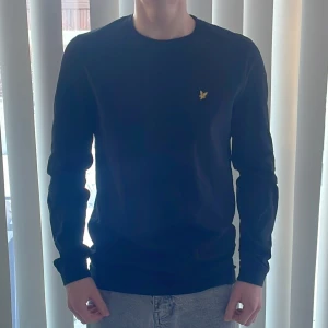 Svart tröja från Lyle & Scott - Säljer en stilren svart långärmad tröja från Lyle & Scott med deras ikoniska gula logga på bröstet. Tröjan har långa ärmar och en klassisk passform. Perfekt för en avslappnad look. Passar ungdomar i åldern 14-15 år ✅
