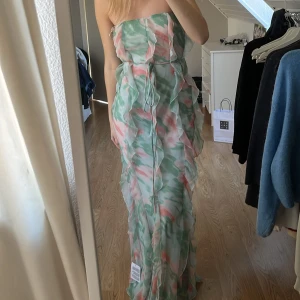 Sommarklänning🩷💚 - Säljer en axelbandslös långklänning med ett vackert grönt och rosa mönster. Klänningen har volangdetaljer och ett knytband i midjan för en smickrande passform. Perfekt för sommarens festligheter!
