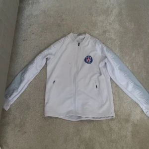 Vit jacka med PSG-logga - Säljer en vit jacka med PSG-logga på bröstet. Jackan har dragkedja framtill och två sidofickor med dragkedjor. Ärmarna har en subtil mönstrad detalj. Perfekt för fotbollsfans som vill visa sitt stöd.