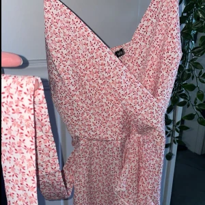 Blommig omlottklänning - Säljer en söt blommig omlottklänning i rosa och vit med volangdetaljer. Klänningen har en v-ringad hals och är ärmlös, perfekt för sommarens varma dagar. Den har en dragkedja i sidan för enkel påklädning och ett knytband i midjan för en fin passform.