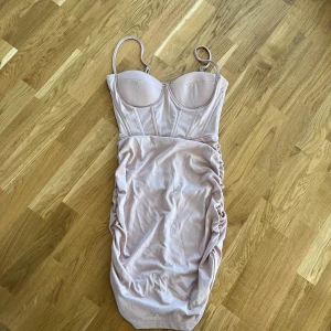 Ljusrosa klänning med rynkdetaljer - Säljer en elegant ljusrosa klänning med smala axelband och rynkdetaljer längs sidorna. Klänningen har en figurnära passform och en stilren design som framhäver formerna. Perfekt för en kväll ute eller en speciell tillställning.