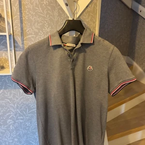 Grå pikétröja från Moncler - Snygg grå pikétröja från Moncler med klassisk krage och knappar. Tröjan har röda och vita detaljer på kragen och ärmsluten samt Monclers logga på bröstet. Perfekt för en stilren look. Passar S/M, köpt på NK.