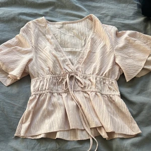 Beige blus med knytband - Säljer en stilren beige blus med v-ringning och knytband i midjan. Blusen har korta ärmar och ett lätt rynkat tyg som ger en fin struktur. Perfekt för en avslappnad men elegant look.