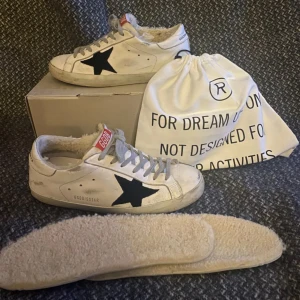 Golden goose - Säljer ett par vita sneakers från Golden Goose med en svart stjärna på sidan. Skorna har en unik design med snörning och en mysig foder inuti. Perfekta för en stilren och bekväm look.