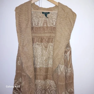 Beige ärmlös kofta från Ralph Lauren - Säljer en snygg beige ärmlös kofta från Ralph Lauren. Den är tillverkad i en blandning av silke, linne och bomull, vilket ger en mjuk och bekväm känsla. Koftan har ett subtilt mönster och en öppen design som gör den perfekt för lager-på-lager-stil. Perfekt för att ge din outfit en elegant touch.