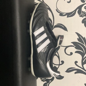 Adidas Copa Mundial fotbollsskor - Klassiska Adidas Copa Mundial fotbollsskor i svart och vitt. Skorna har en tidlös design med tre vita ränder på sidan och är tillverkade i skinn för optimal komfort och hållbarhet. Perfekta för fotbollsplanen med sina dobbar för bra grepp.