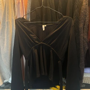 Svart ribbad topp från Nelly - Säljer en stilren svart ribbad topp från Nelly med lång ärm och en unik knytning framtill. Helt oanvänd och har prislappen kvar 