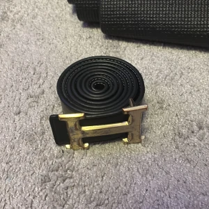 Svart bälte med guldfärgat H spänne  - Snyggt svart bälte med ett iögonfallande guldfärgat H-spänne. Perfekt för att ge din outfit en stilren touch. Bältet är tillverkat i ett slätt material och har en klassisk design.