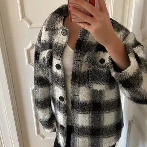 Rutig teddyjacka i svart och vit - Supermysig teddyjacka i svart och vit med rutigt mönster. Perfekt för kyliga dagar med sina långa ärmar och knappar framtill. Jackan har en skön och fluffig känsla, vilket gör den idealisk för höst och vinter. Två praktiska bröstfickor med knappar för extra stil och funktion. Endast använd en gång och är därav i nytt skick.