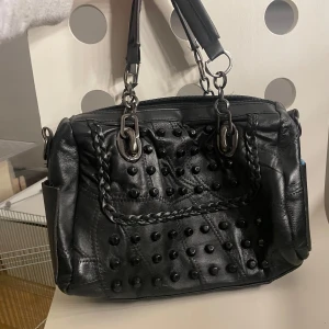 Svart handväska med nitar - Snygg svart handväska i skinn med coola nitar på framsidan. Väskan har flätade detaljer och robusta handtag med metallringar. Perfekt för en edgy look och rymmer allt du behöver för en dag på stan.