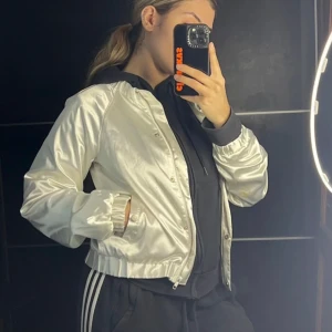 Vit bomberjacka - Snygg vit bomberjacka med dragkedja och ribbade muddar. Perfekt för en cool och avslappnad stil. Jackan har en glansig finish och normal passform.