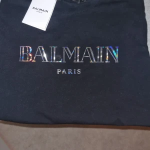 Svart t-shirt från Balmain - Snygg svart t-shirt från Balmain med holografiskt tryck av märkesnamnet på framsidan. Perfekt för en stilren look.