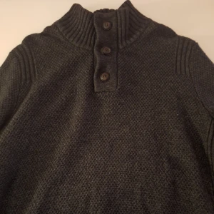 stickad tröja - Säljer en stilren mörkgrå stickad tröja med ribbad krage och tre knappar framtill. Perfekt för kyliga dagar och ger en klassisk look. Passar bra till både jeans och chinos. Storlek l men passar också för storlek m. Pris ej hugget I sten