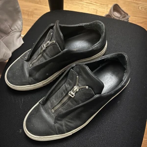 Svarta sneakers med dragkedja - Snygga svarta sneakers från Santoni med dragkedja framtill. Skorna har en stilren design med vit sula som kontrasterar mot det svarta. Perfekta för en avslappnad men trendig look.