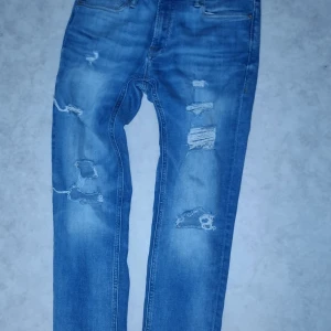 Blå skinny jeans från Jack & Jones - Säljer ett par blå skinny jeans från Jack & Jones, modell Liam. De har en sliten look med flera revor och slitningar för en trendig stil. Perfekta för dig som gillar en tight passform och modern design.