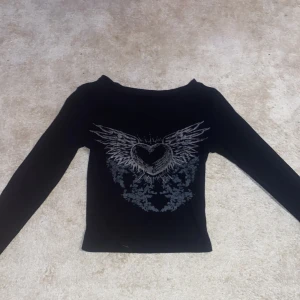 Alt y2k lång ärmad tröja  - Snygg svart långärmad topp med ett coolt hjärtmönster och vingar på framsidan. Perfekt för en edgy look. Toppen har en tight passform och är croppad för en modern stil.