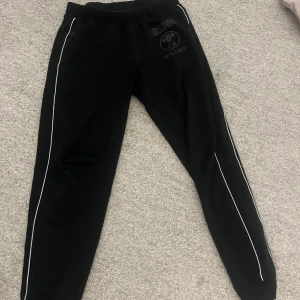 Svarta mjukisbyxor från Moschino - Snygga svarta mjukisbyxor från Moschino med vita detaljer längs benen. Perfekta för en avslappnad stil med en elastisk midja för extra komfort. Moschino-loggan syns tydligt på framsidan. Köpt för 1600 från kidsbrandstore förra månaden. Dom är som helt nya 