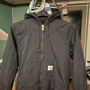 Svart jacka från Carhartt - Säljer en svart jacka från Carhartt med huva och dragkedja. Jackan har en klassisk design med Carhartt-logga på framsidan. Perfekt för kyligare dagar med Thinsulate-isolering för extra värme.