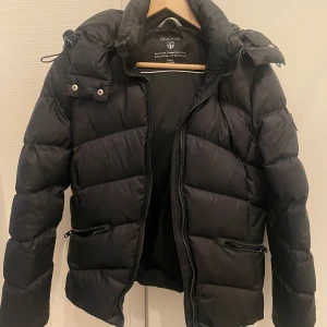 Svart dunjacka från GANT - Säljer en svart dunjacka från GANT i mycket bra skick. Jackan har en avtagbar huva och dragkedja framtill. Inga skador på jackan dock trög dragkedja.  Ny pris 3500kr