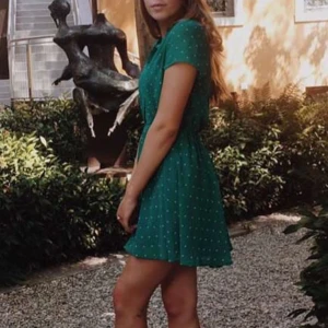 Grön kortklänning med prickar - Charmig grön kortklänning med vita prickar och korta ärmar. Perfekt för en somrig dag med sin lätta och luftiga design. Klänningen har söta knappar framtill som ger en extra touch av stil.