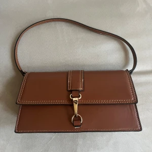 Brun axelväska i skinn - Snygg brun axelväska i skinn med stilren design. Väskan har en elegant gulddetalj på framsidan och en praktisk axelrem. Perfekt för både vardag och fest! Inköpt på Mango förra året i Los Angeles. I nyskick. 