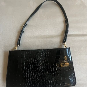 Svart axelväska i skinn från And other stories! - Snygg svart axelväska i skinn med krokodilpräglat mönster. Väskan har en justerbar axelrem och två dragkedjor upptill för säker förvaring. Inköpt för cirka 2-3 år sedan från And other stories i Göteborg. 