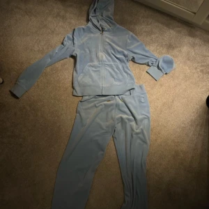 Blå velour hoodie och byxor från Juicy Couture - Säljer en mjuk och bekväm blå velour hoodie och matchande byxor från Juicy Couture. Hoodien har dragkedja och fickor framtill, medan byxorna har en elastisk midja med snörning. Perfekt för en avslappnad stil.