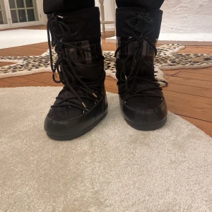 Svarta Moon Boots med snörning - Snygga och varma svarta Moon Boots med hög design och snörning. Perfekta för kalla vinterdagar. De har en glansig yta och en robust sula för extra grepp.