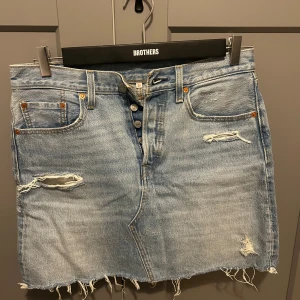 Blå jeanskjol från Levi's - Snygg blå jeanskjol från Levi's med slitna detaljer och fransig nederkant. Kjolen har storlek 30 enligt Levi’s och skulle säga det är ungefär M/L