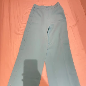 Turkosa byxor från FB Sister - Snygga stark turkosa byxor från FB Sister med knappar längs sidorna. Byxorna har en elastisk midja för extra komfort och en loose passform. Perfekta för en avslappnad men stilig stil.