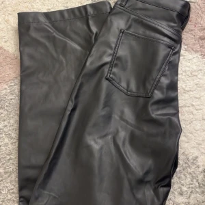 Svarta byxor i syntetmaterial - Snygga svarta byxor i syntetmaterial med hög midja och raka ben. Perfekta för en trendig look. Byxorna har fickor både fram och bak samt en klassisk dragkedja och knapp framtill.