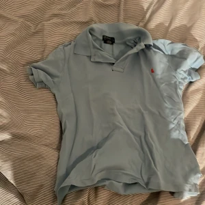 Ljusblå pikétröja från Ralph Lauren - Säljer en ljusblå pikétröja från Ralph Lauren i bra skick. Den har en klassisk krage och korta ärmar, med det ikoniska röda broderade logotypen på bröstet. Perfekt för både vardag och lite finare tillfällen. Skön och stilren! 💙