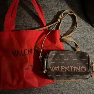 Valentino axelväska och shoppingväska - Säljer en snygg Valentino axelväska i brunt med logotypmönster och dragkedja, perfekt för vardagsbruk. Medföljer en röd shoppingväska med Valentino-tryck, idealisk för shoppingturer. Båda väskorna är i mycket bra skick och ger en lyxig touch till din outfit.