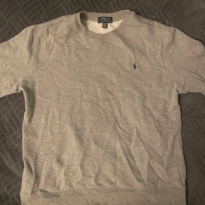 Grå Polo Ralph Lauren sweatshirt - Säljer en klassisk grå sweatshirt från Polo Ralph Lauren i bra skick. Tröjan har en liten broderad logga på bröstet och är perfekt för både vardag och avslappnade tillfällen. Den är mjuk och bekväm med normal passform. Perfekt för höst och vår! Har ett litet hål på vänster armen som syns på andra bilden.
