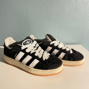 Adidas Campus 00 - Säljer ett par svarta Adidas Campus 00 med vita ränder och snören. De har en gummisula i beige. Själva skorna är i fint skick, det är endast snörena som är lite smutsiga. Kom gärna med prisförslag eller frågor:)