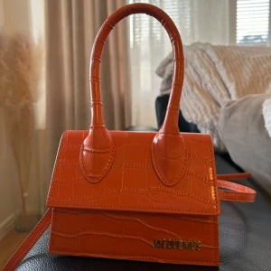 Orange handväska från Jacquemus - Snygg orange handväska från Jacquemus i krokodilpräglat skinn. Väskan har ett stilrent handtag och en justerbar axelrem. Perfekt för att ge en färgklick till din outfit!