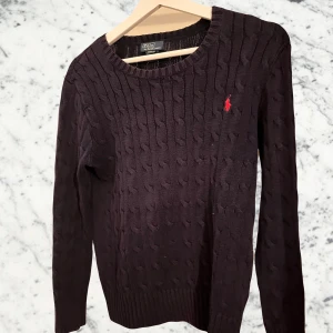 Marinblå kabel stickad tröja från Ralph lauren - Säljer en snygg blå stickad tröja från Polo Ralph Lauren. Den har ett klassiskt kabelstickat mönster och en liten röd logga på bröstet. Perfekt för både höst och vinter, med långa ärmar och ribbade kanter. Passar till både vardag och fest!