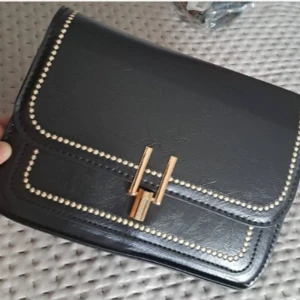 Svart axelväska med gulddetaljer - Snygg svart axelväska i syntetmaterial med eleganta gulddetaljer. Väskan har en stilren design med en dekorativ guldfärgad låsdetalj på framsidan. Perfekt för både vardag och fest!