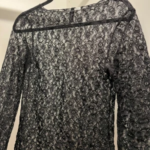 Svart spets topp  - Elegant svart spets topp med långärmad design. Toppen har ett vackert blommigt mönster och en transparent look som ger en sofistikerad stil. Perfekt för speciella tillfällen.