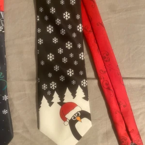 Julslipsar med motiv - Säljer ett set med tre julslipsar. En röd slips med julgran och dekorationer, en svart slips med snöflingor och en pingvin i tomteluva, samt en slips med tomten på motorcykel. Perfekt för att sprida julstämning!!