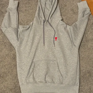 Grå hoodie från Ami Paris - Säljer en stilren grå hoodie från Ami med en liten röd logga på bröstet. I storlek L men kan även passa M.Den har en klassisk känguruficka och justerbar huva med snören. Perfekt för en avslappnad look. Tar även byten. Hör av er vid fler frågor 