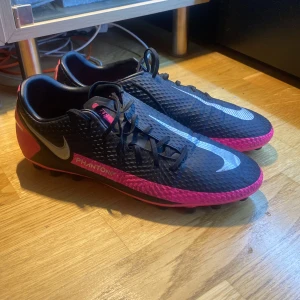 Nike Phantom fotbollsskor - Snygga Nike Phantom fotbollsskor i svart och rosa med snörning. Skorna har ett väldigt bra skick förutom en liten defekt på vänsra sula som man ser på bild 2, men det är inget märkvärdigt! Köpte skorna för ca 1500kr. Hör av er vid frågor och funderingar😃