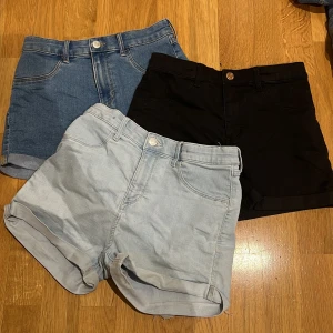 Tre par shorts i olika färger - Säljer tre par shorts i olika färger. Ett par ljusblå jeansshorts med upprullade ben, ett par svarta shorts och ett par mörkblå jeansshorts. Perfekta för sommaren och enkla att matcha med olika outfits.