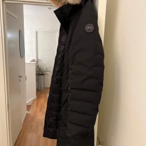 Svart dunjacka från Canada Goose - Säljer en svart vinterjacka från Canada Goose i mycket bra skick. Jackan har en pälskantad huva och är perfekt för kalla vinterdagar. Den är långärmad och har en snygg, stilren design med en broderad logga på ärmen. Perfekt för både vardag och vinteräventyr!