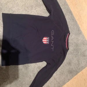 Mörkblå tröja från GANT - Säljer en snygg mörkblå tröja från GANT med ett coolt emblem på bröstet. Tröjan har långa ärmar och är perfekt för både vardag och lite mer avslappnade tillfällen. Den är i bra skick och redo för en ny ägare!